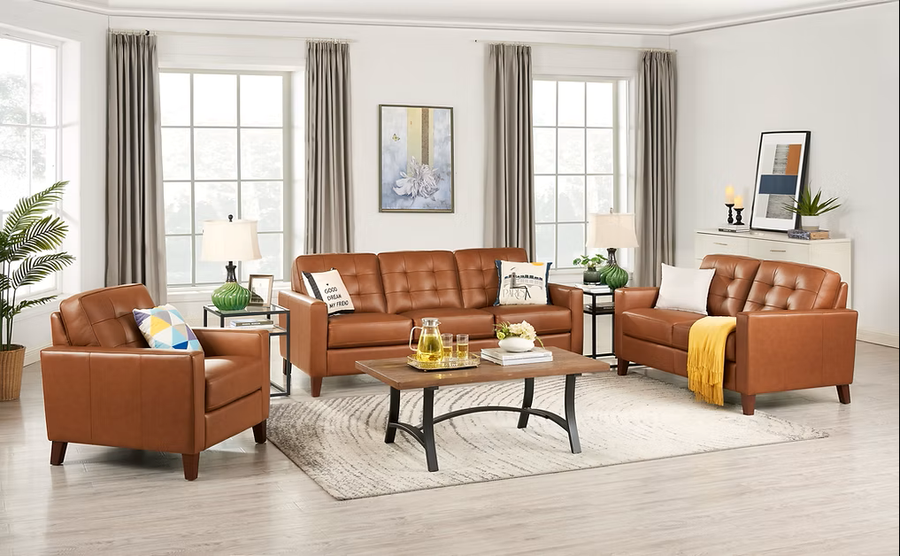Three-piece Aiden Collection in warm nutmeg leather upholstery, featuring a three-seat sofa, loveseat, and armchair. Each piece has square track arms, tufted back cushions, and tapered wood legs. Arranged around a rectangular coffee table in a bright, modern living room.