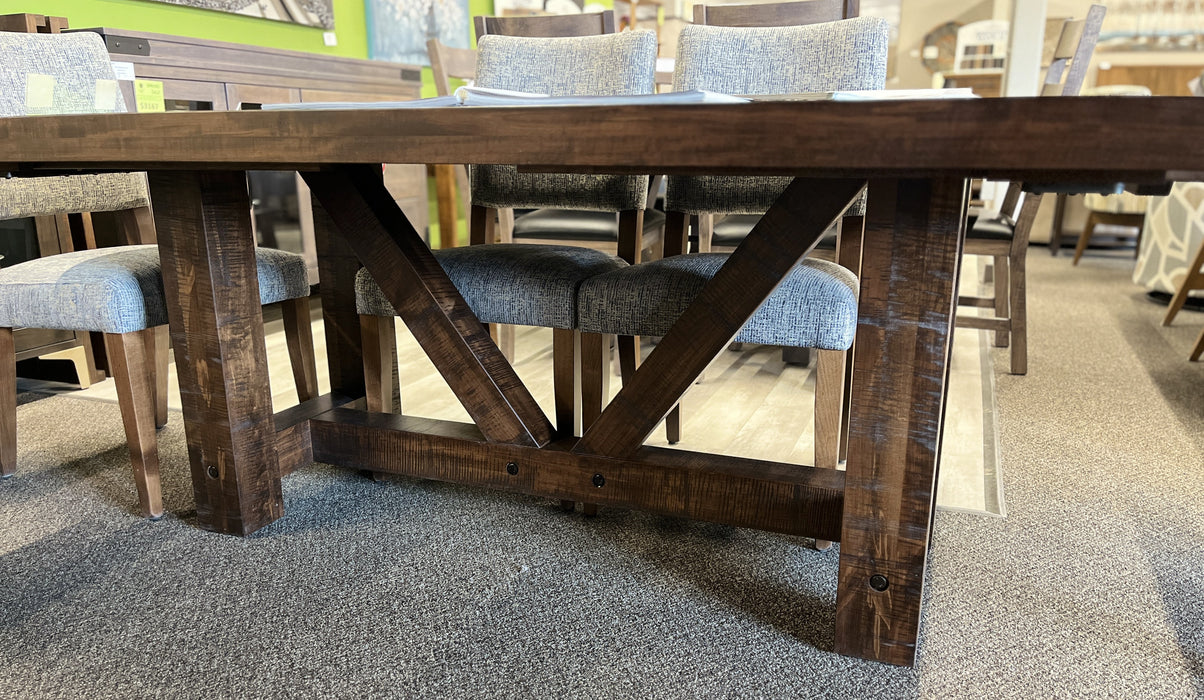 Close-up of Balmoral solid wormy maple dining table in Mocha stain, showing its rough-sawn wood texture, substantial square legs, angled trestle supports and dark accent fasteners, with a few Tampa dining chairs visible behind.