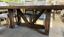 Close-up of Balmoral solid wormy maple dining table in Mocha stain, showing its rough-sawn wood texture, substantial square legs, angled trestle supports and dark accent fasteners, with a few Tampa dining chairs visible behind.