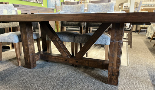 Close-up of Balmoral solid wormy maple dining table in Mocha stain, showing its rough-sawn wood texture, substantial square legs, angled trestle supports and dark accent fasteners, with a few Tampa dining chairs visible behind.